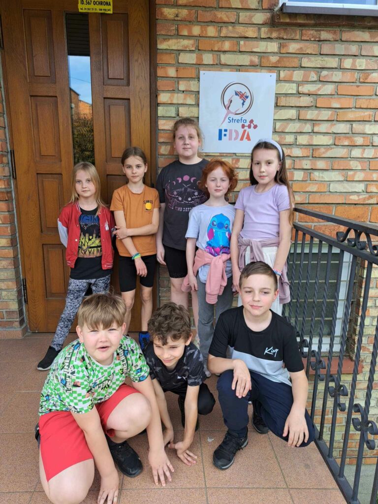 Zdjęcie przedstawia 8 dzieci stojących przed budynkiem, w którym odbywają się zajęcia fitness jumping. 